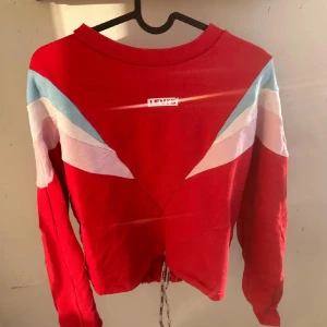 Röd Levi's sweatshirt med snörning - röd sweatshirt från Levi's med färgglada detaljer i ljusblått, vitt och rosa på axlarna. Tröjan har lång ärm, rund hals och snörning som är stretch där nere som man kan dra åt. Den är croopad lös passform. Den är S o passar M oxå