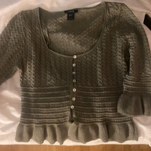 Grön kofta med volang från Lindex - Supersöt grön stickad kofta från Lindex med dekorativa knappar framtill och volangdetaljer vid nederkant och ärmslut. Koftan har ett snyggt hålmönster och trekvartsärmar.
