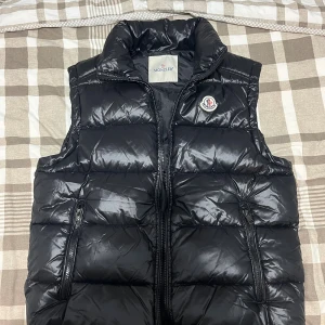 Svart dunväst från Moncler - Svart, glansig dunväst från Moncler Storlek S med hög krage och två dragkedjefickor framtill. Klassisk Moncler-logga på bröstet och quiltad design. Perfekt för lager-på-lager och snygg till streetwear.