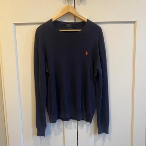 Marinblå tröja från Polo Ralph Lauren - Snygg marinblå tröja från Polo Ralph Lauren i mjuk pima bomull. Klassisk rund hals och långärmad modell med den ikoniska orangea logon broderad på bröstet. Perfekt för dig som gillar stilrena och tidlösa plagg.