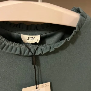 Grön blus med volang från JDY - Supersöt grön blus från JDY med volangdetaljer vid halsringning och ärmslut. Blusen har korta ärmar och en rak passform som känns både enkel och trendig. Perfekt att styla med jeans eller kjol för en fräsch look.
