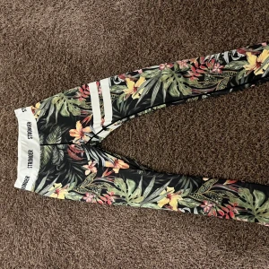 Blommiga leggings från Stronger - Färgglada leggings från Stronger med tropiskt blommönster i grönt, gult, rött och rosa. De har en vit resår med logga och vita ränder på ena benet. Perfekta för träning och riktigt sköna att ha på sig. Dom är lite noppriga Ibai men syns inte så väl 