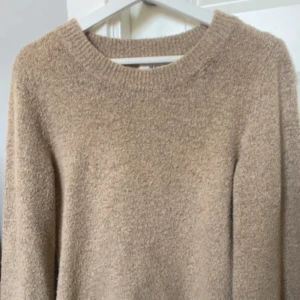 Beige stickad tröja med rund hals - Mysig beige stickad tröja med rund halsringning och långa ärmar. Tröjan har en fluffig och mjuk struktur som gör den perfekt för kyligare dagar. Enkel och stilren design som är lätt att matcha med olika outfits. Nopprig 