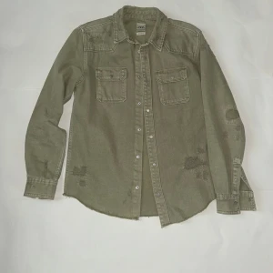 Olivgrön overshirt från Zara - Olivgrön overshirt från Zara med rå kant nedtill och slitna detaljer på axlar, ärmar och framstycke. Två bröstfickor med knappar och klassisk krage. Långärmad och tillverkad i kraftig bomull som ger en cool och avslappnad look.