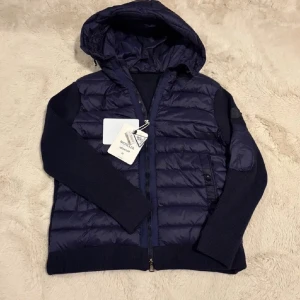 Mörkblå pufferjacka från Moncler - Snygg mörkblå pufferjacka från Moncler med huva och stickade ärmar. Jackan har quiltad framsida, dragkedja och en ficka med zip på ärmen. Materialmix av polyester och stickat ger en cool look. Moncler-logga på ärmen för extra stilpoäng.