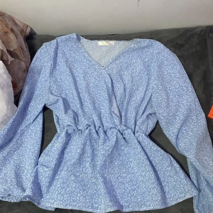 Blommig blå omlottblus från By-L - Säljer en söt blå blus från By-L Studio med vitt blommigt mönster. Blusen har omlottdesign, v-ringning och långa ärmar. Midjan är markerad med en lätt rynkning för en snygg siluett. Perfekt för dig som gillar romantiska och fräscha looks!