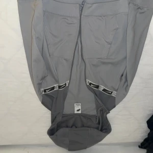 Grå Nike hoodie med dragkedja - Snygg grå hoodie från Nike med hel dragkedja och huva. Tröjan har två fickor framtill och coola svarta och vita Nike-detaljer vid fickorna. Perfekt för chill eller träning, med en modern och sportig look.