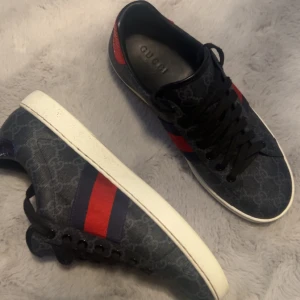 Gucci sneakers med GG-mönster - Snygga svarta Gucci sneakers med klassiskt GG-monogram i tyg och skinn. Skorna har röda och mörkblå ränder på sidorna, svart snörning och vit sula. Insidan är dekorerad med GG-logga och detaljer i läder. Kommer med originalkartong.