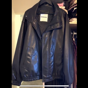 Svart skinnjacka från Bershka - Säljer en svart skinnjacka från Bershka med klassisk krage och dragkedja framtill. Jackan har två snedställda fickor och elastiska muddar vid ärmslut och nederkant. Oversized passform för en cool och avslappnad stil.
