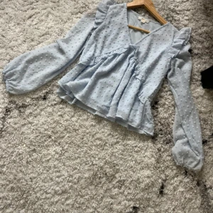 Ljusblå blus med volanger och prickar - Supersöt ljusblå blus med volangdetaljer och diskret prickigt mönster. Blusen har v-ringning, långa ärmar med puff och lätt transparent tyg. Perfekt för dig som gillar romantisk och feminin stil!