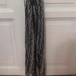 Glittrig Missoni halsduk i zigzag-mönster - Snygg halsduk från Missoni med klassiskt zigzag-mönster i svart, grått och vitt. Halsduken har glittriga inslag och fransar i ändarna. Tillverkad i en mix av ull, akryl, viskos och polyester. Perfekt accessoar för att lyfta din outfit.