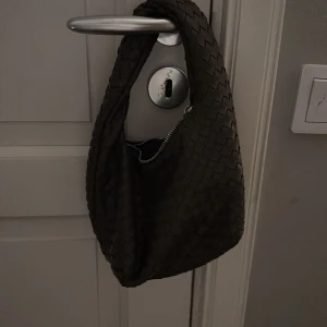 Svart flätad axelväska i skinn - Snygg  axelväska med flätad design och dragkedja upptill. Väskan har en mjuk, rundad form och är tillverkad i skinn, vilket ger en lyxig känsla. Perfekt storlek för att bära med sig det viktigaste och passar till många olika stilar.