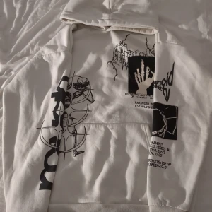 Vit hoodie med grafiskt tryck Black Squad - Vit hoodie från Black Squad i relaxed fit med huva och coola grafiska tryck i svart på både fram- och baksida. Trycken har motiv som hand, skalle, måne och text. Tröjan har lång ärm och är tillverkad i mjuk bomull, perfekt för dig som gillar streetwear och unika detaljer.