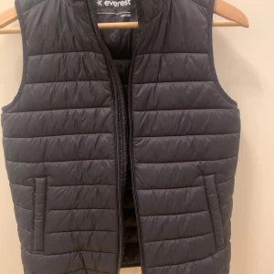 Svart täckväst från Everest, Stadium - Svart quiltad täckväst från Everest, Stadium, med dragkedja framtill och två fickor. Västen har en stilren, rak passform och är tillverkad i nylon med polyesterfyllning. Perfekt för lager-på-lager och funkar till många olika stilar.