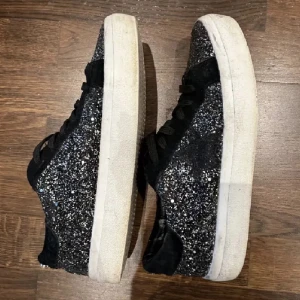 Svarta glittriga sneakers Steve Madden - Svarta sneakers från Steve Madden med glittrig yta och svarta mockadetaljer. Skorna har snörning, vit platt sula och en stjärndetalj på sidan. Perfekta för dig som vill sticka ut med lite extra bling i vardagen. Passar 39-40. Köpt för 700 säljer för 200