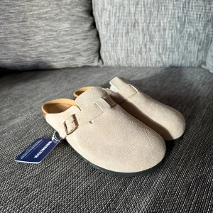 Beige Birkenstock Boston i mocka - Säljer ett par Birkenstock Boston i beige mocka med klassisk rund tå och justerbart spänne i metall. Skorna har en platt sula i kork och svart gummi, perfekt för chill vardagsstil. Superbekväma och tidlösa slip-ins som funkar året runt.