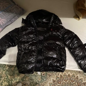 Svart pufferjacka från Polo Ralph Lauren - Säljer en svart, glansig pufferjacka från Polo Ralph Lauren i storlek M. Jackan har huva, dragkedja framtill och två stora fickor. Klassisk röd Polo-logga broderad på bröstet. Perfekt för kalla dagar och har en riktigt schysst streetstil.