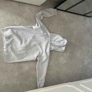 Grå hoodie från BVALDI med paljettdetaljer - Snygg ljusgrå hoodie från BVALDI med dragkedja och huva. Framsidan är dekorerad med coola paljettdetaljer i form av händer och text. Perfekt för dig som vill sticka ut med en unik och bekväm stil.