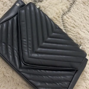 Svart quiltad axelväska med kedja - Snygg svart axelväska med quiltad design och silverfärgad kedja som axelrem. Väskan har ett geometriskt mönster med V-formade sömmar och är tillverkad i skinnimitation. Perfekt storlek för att få plats med allt viktigt och ger en cool vibe till din outfit.