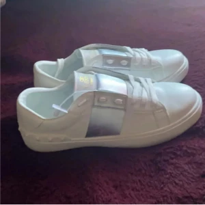 Valentino Garavani vita sneakers - Säljer ett par snygga vita Valentino Garavani sneakers med silvriga detaljer på sidan och klassisk snörning. Skorna har en platt sula med mönstrad undersida och är tillverkade i skinn. Perfekt för dig som gillar exklusiva och stilrena sneakers med lyxig känsla.