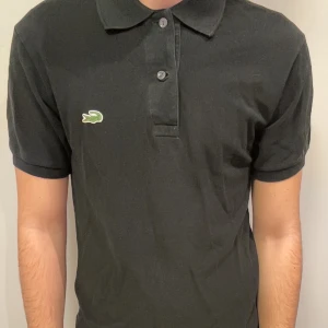 Svart pikétröja från Lacoste - Svart pikétröja från Lacoste med klassisk krage och korta ärmar. Tröjan har två knappar framtill och den ikoniska gröna krokodilloggan på bröstet. Tillverkad i mjuk bomull som är skön mot huden och passar perfekt till en chill stil. Hör av er för prisförslag, går alltid att finslipa priset lite🤝🤩