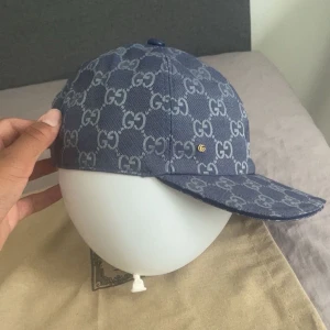 Blå keps från Gucci med GG-mönster - Säljer en blå keps från Gucci med ikoniskt GG-mönster över hela kepsen. Kepsen har böjd skärm, justerbar rem bak och en liten guldfärgad GG-detalj på sidan. Tillverkad i slitstarkt tyg och levereras med originalförpackning och tags.