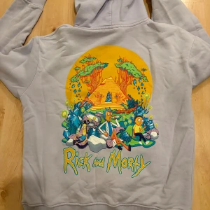 Rick and Morty ljusgrå/lila hoodie - Säljer en ljusgrå/lilahoodie med färgglad Rick and Morty-tryck både fram och bak. Tröjan har huva, magficka och ribbade muddar. Perfekt för dig som älskar serien och vill sticka ut med ett coolt motiv.