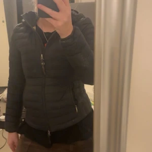 Svart dunjacka från Parajumpers - Svart dunjacka från Parajumpers med quiltad design och dragkedja framtill. Jackan har huva, sidofickor med dragkedja och ljus insida. Klassisk patch på axeln. Perfekt för kalla dagar och har en snygg, slim passform. den är äkta såklart. några få hål som visas på bilden därför priset🤍. köptes för 4500kr