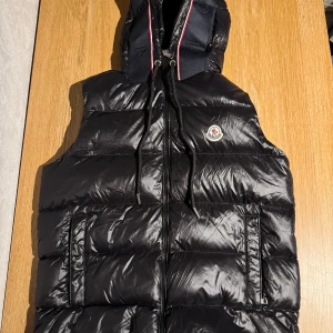 Svart dunväst från Moncler med huva - glansig dunväst från Moncler med huva och dragkedja framtill. Västen har två fickor med dragkedja, broderad Moncler-logga på bröstet och snygga detaljer med röd, vit och blå kant på huvan. Nfc tagg fungerar.