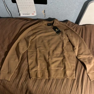 Beige sweatshirt från Lyle & Scott - Snygg beige sweatshirt från Lyle & Scott med klassisk rund hals och ribbade muddar. Tröjan har ett broderat gult fågelmärke på bröstet och är tillverkad i mjuk bomull. Perfekt för chill dagar och enkel att matcha med jeans eller joggers.