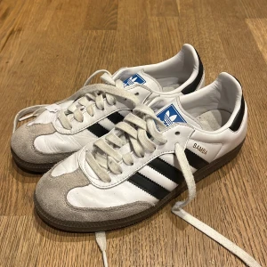 Adidas Samba vita och svarta sneakers - Klassiska Adidas Samba sneakers i vitt skinn med svarta ränder och detaljer. Skorna har grå mocka vid tån, bruna gummisulor och snörning framtill. Perfekta för dig som gillar retrostil och sportig vibe.