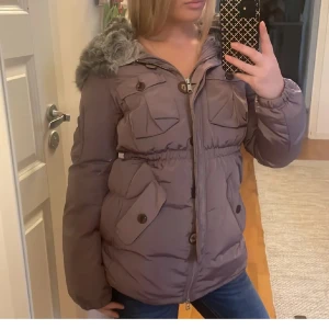 pufferjacka Odd Molly - Säljer en brun lila  pufferjacka från Odd Molly med fluffig fuskpäls på huvan, flera fickor med knappar och dragkedja framtill. Jackan har markerad midja och långa ärmar, perfekt för kalla dagar. Snygg och mysig stil som sticker ut.
