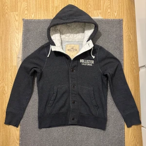 Grå Vintage Hollister Caridgan Med Knappar - Väldigt sällsynt och vintage Hollister kofta/caridgan. Tröjan har stora knappar framtill, dragsko i huvan och två fickor. Denna koftan är i mycket bra skick! Etiketten säger Storlek L men passar M, 180-187. Längd ≈ 65 cm, Bredd armhåla - armhåla ≈57cm. Modell 174-175, 70kg