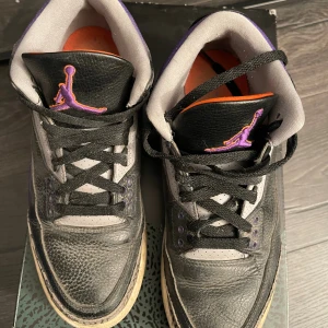 Nike Air Jordan 3 Court Purple - Nike Air Jordan 3 Court Purple med svart och grått skinn, lila och orange detaljer samt klassiskt elephant print runt hälen. Skorna har svart snörning, orange innersula och lila Jumpman-logga på plösen och hälen. Perfekt för dig som gillar unika sneakers med retrovibbar. Köpta på sneakers n stuff
