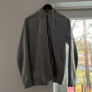 Grå hoodie från Moncler med dragkedja - Snygg grå hoodie från Moncler med huva och vita snören. Dragkedja framtill med röd och blå kant, samt Moncler-logga på bröstet. Långärmad med diskret röd och blå rand på ärmen. Perfekt för chill dagar och streetstyle.