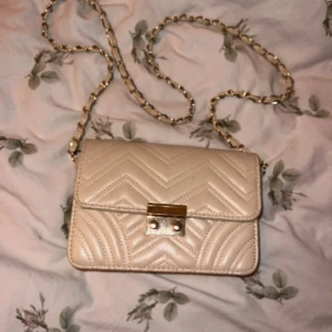 Beige axelväska med kedja - Snygg beige axelväska med quiltat zigzag-mönster och guldfärgad kedja som axelrem, väskan är från h&m i använt skick men fortfarande väldigt fin.