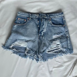 Blå slitna jeansshorts från Signature8 - Säljer ett par blå denimshorts från Signature8 med hög midja, slitna detaljer och fransig kant. Shortsen har knappar framtill, klassiska fickor och en cool, avslappnad vibe. Perfekta för sommardagar när du vill ha en edgy look.