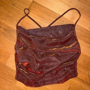 Glittrig vinrött linne med  öppen rygg - Säljer en glittrig vinröd linnetopp med tunna axelband som korsas i ryggen. Toppen har en snygg draperad front och skimrar i både rött och guld beroende på ljuset. Linnet är använt fåtal gånger och är i fint skick det är från SHEIN och är i storlek M