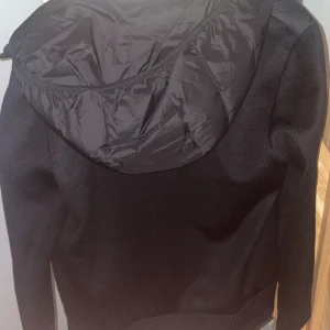 Svart Moncler cardigan med huva - Svart pufferjacka från Moncler med stickade ärmar och quiltad front. Jackan har en snygg huva i blankt material och dragkedja framtill med Moncler-logga längs kanten. Perfekt för dig som vill ha en stilren och modern look. Storlek m passar även s väldigt bra. Pris kan diskuteras 