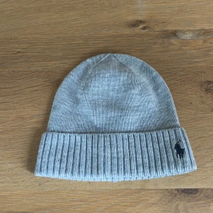 Grå ullmössa från Polo Ralph Lauren - Snygg grå mössa från Polo Ralph Lauren i 100% ull. Klassisk ribbstickad kant och broderad mörkblå Polo-logga framtill. Enkel och stilren design som passar perfekt till kalla dagar. Mössan är mjuk och har en neutral grå färg som är lätt att matcha.