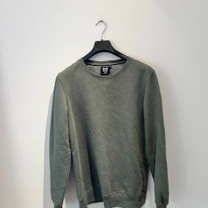 Grön sweatshirt från Revolution - En enkel och stilren grön sweatshirt från Revolution i storlek XL. Tröjan har rund hals, ribbade muddar vid ärmslut och nederkant samt en liten läderdetalj i nacken. Perfekt för chill dagar och passar till jeans eller joggers. Hör av er vid frågor