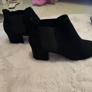 Svarta chelsea boots i mocka - Snygga svarta chelsea boots i mocka med låg klack och rund tå. Skorna har elastiska paneler på sidorna för enkel påtagning. De är i gott skick och har inga defekter samt en klack på 5cm