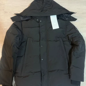 Canada Goose  - Säljer en Canada Goose jacka svart logo med huva och diskret logga på ärmen. Jackan har raka linjer, stora fickor och stängs med både dragkedja och knappar. Perfekt för kalla dagar och riktigt snygg till streetwear. Påsen och pälsen följer med.