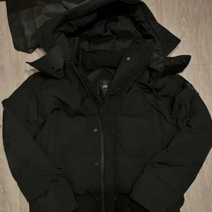 Canada Goose - Säljer en Canada Goose jacka med svart logo med klassisk patch på ärmen. Jackan har huva, dragkedja och knappar framtill samt quiltad design. Perfekt för kalla dagar och riktigt snygg streetstil. Påsen och pälsen följer med.