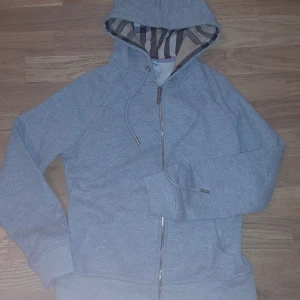 Grå hoodie från Burberry med dragkedja - Snygg grå hoodie från Burberry i storlek S med klassiskt rutigt foder i huvan. Tröjan har dragkedja framtill, snörning vid huvan och ribbade muddar. Perfekt för dig som gillar stilrena och exklusiva streetwear-plagg. Och den är helt ny bätte fin o varm för vinter 