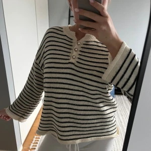 Randig stickad tröja med pärlknappar - Superfin stickad tröja i vitt och svart med smala ränder och tre snygga pärlknappar framtill. Tröjan har lång ärm, rund hals och en loose fit som ger en chill vibe. Perfekt för lager-på-lager under höst och vinter.