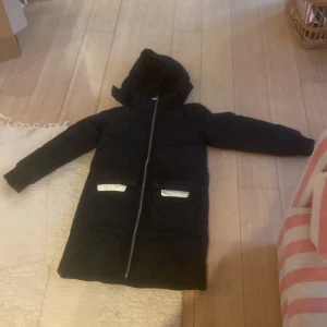 Svart lång pufferjacka Everest - Svart lång pufferjacka från Everest med huva och två fickor med vita detaljer. Jackan har hel dragkedja framtill och är vadderad för extra värme. Perfekt för kalla dagar och har en stilren look som funkar till det mesta.