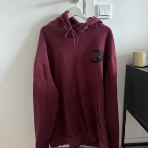 Gasp hoodie - Säljer en vinröd hoodie med svart tryck på bröstet. Tröjan har huva med snörning och en stor magficka. Oversized passform och långa ärmar, perfekt för en avslappnad stil.