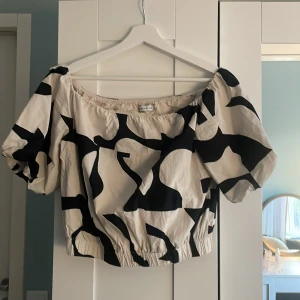 Svartvit offshoulder blus - Trendig offshoulder blus från Stockholm LM med svartvitt abstrakt mönster. Blusen har puffiga korta ärmar och resår i nederkant för en croppad passform. Perfekt till jeans eller kjol för en cool look.