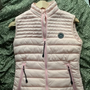 Ljusrosa dunväst från Marine XS - Supersöt ljusrosa dunväst från Marine i storlek XS. Västen har quiltad design, dragkedja framtill och två fickor med dragkedja. Perfekt för lager-på-lager. Finns även fickor på insidan av jackan! Aldrig använts 